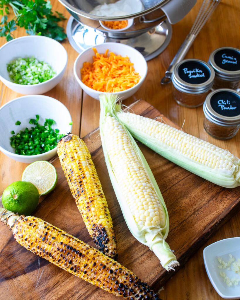 A cutting board with cobs of corn and other ingredients near by.