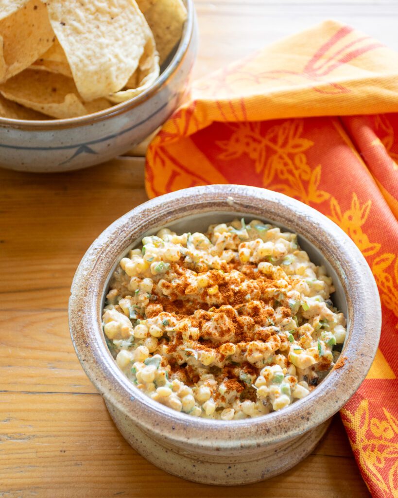 A bowl of corn dip with an orange kitchen towel and some tortilla chips near by.