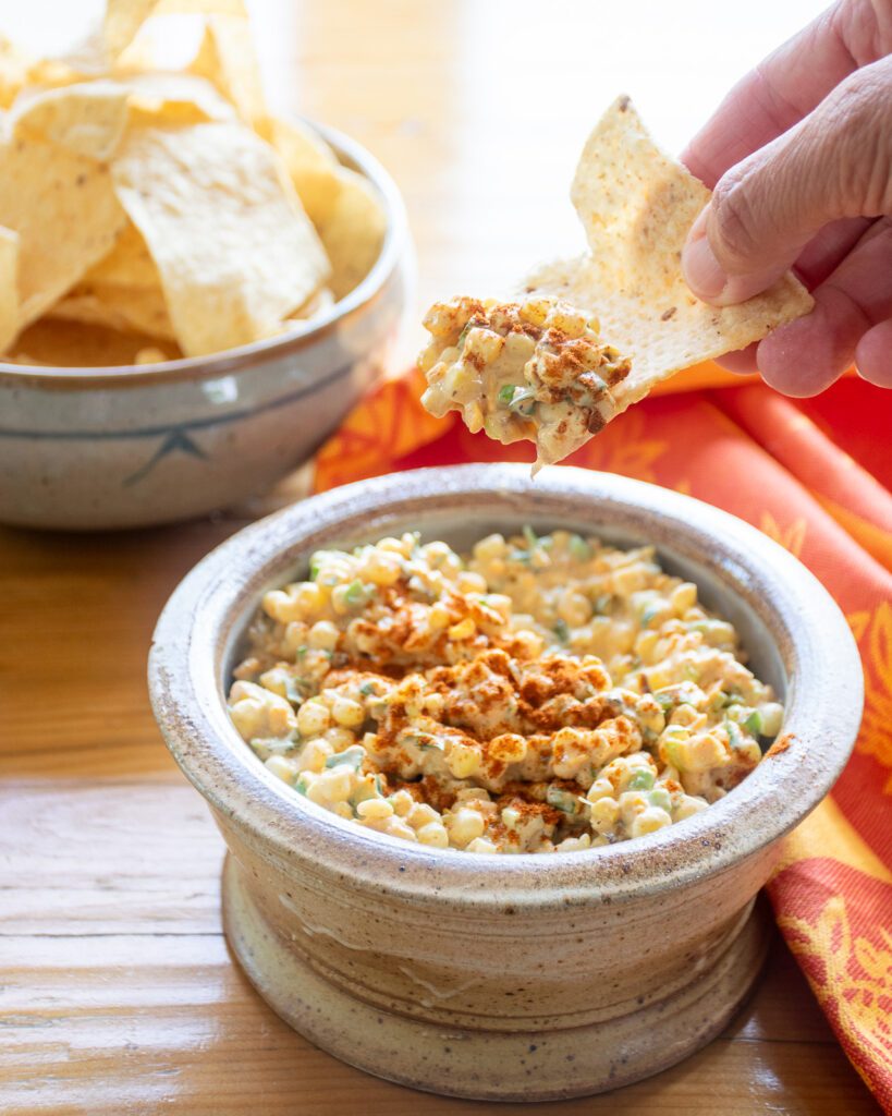 A hand scooping corn dip onto a tortilla chip out of a bowl with an orange kitchen towel near by.