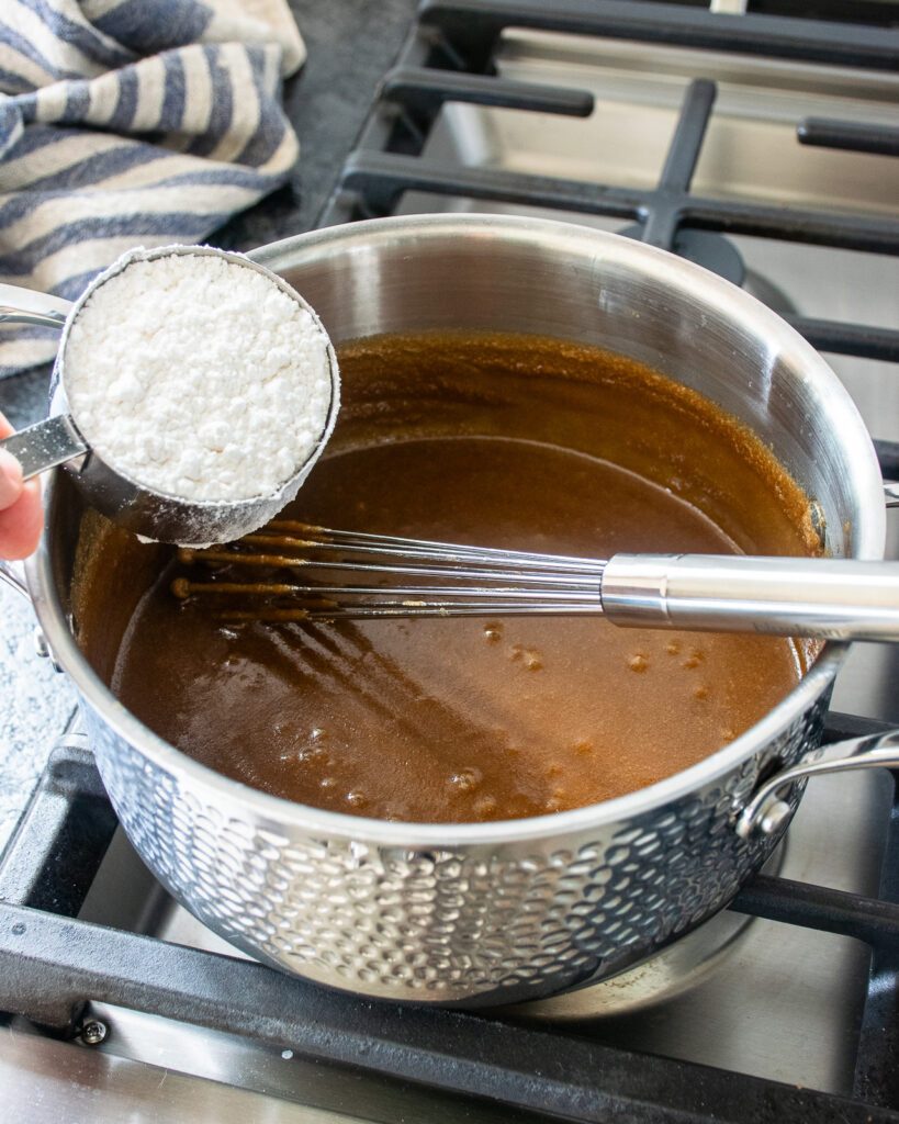 A measuring cup of icing sugar held over a saucepan of brown sugar caramel with a whisk sticking out of it.