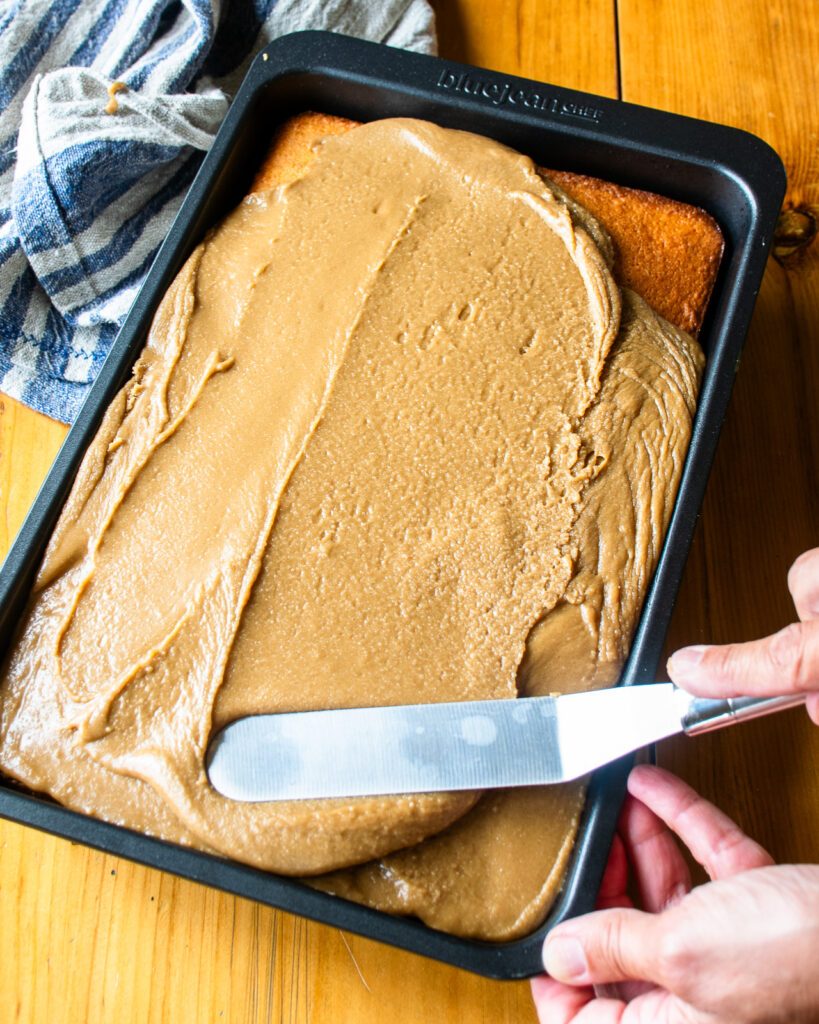 An offset spatula spreading brown sugar icing over hot milk cake.