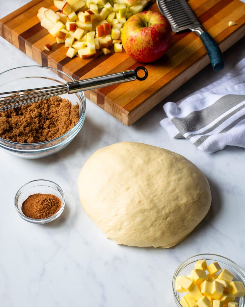 A ball of cinnamon roll dough on a counter with brown sugar, cinnamon, butter and diced apples near by.