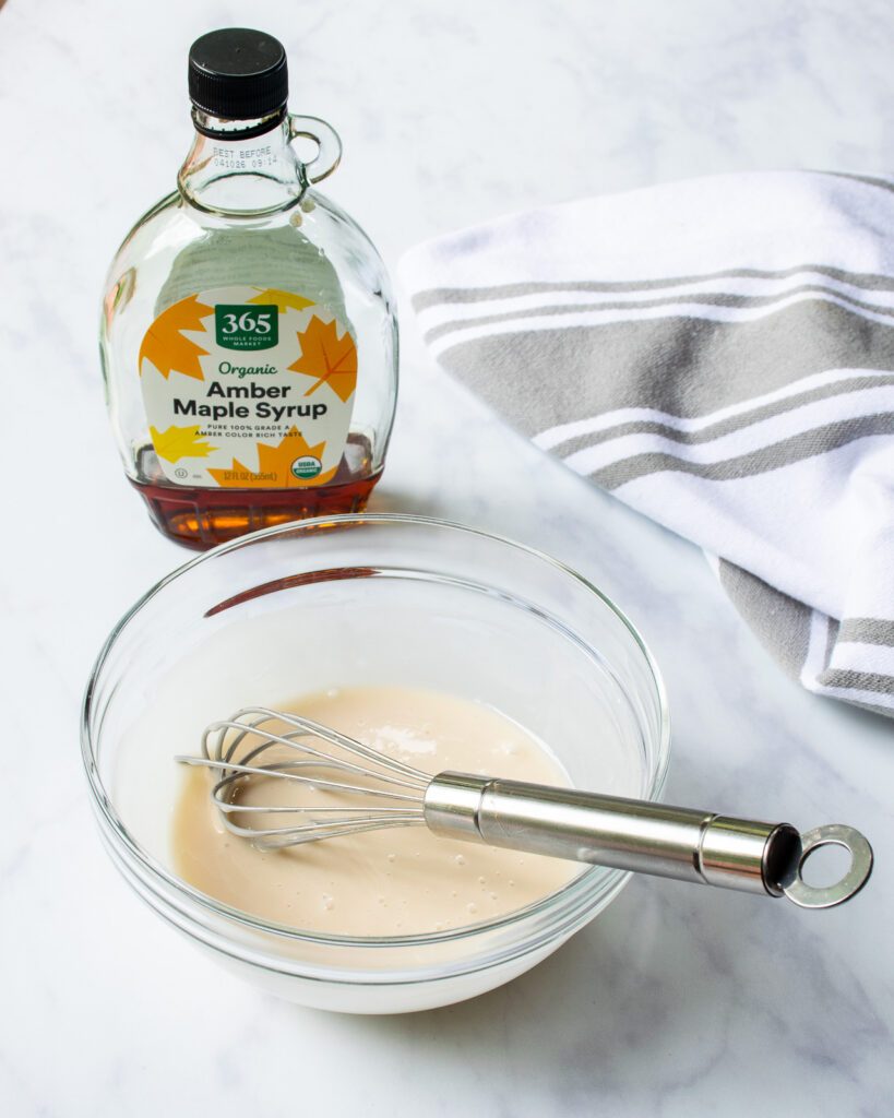 A bowl of maple icing with a bottle of maple syrup in the background.