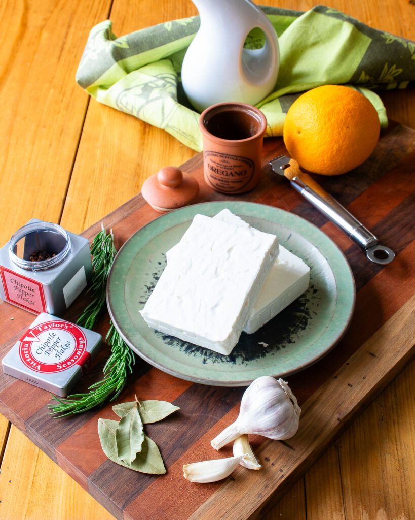 Ingredients for marinated feta cheese on a cutting board - orange, pepper flakes, garlic, bay leaves, rosemary and oregano.