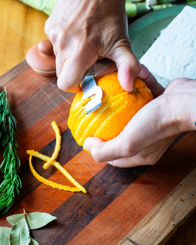 Hands removing orange zest with a channel knife.