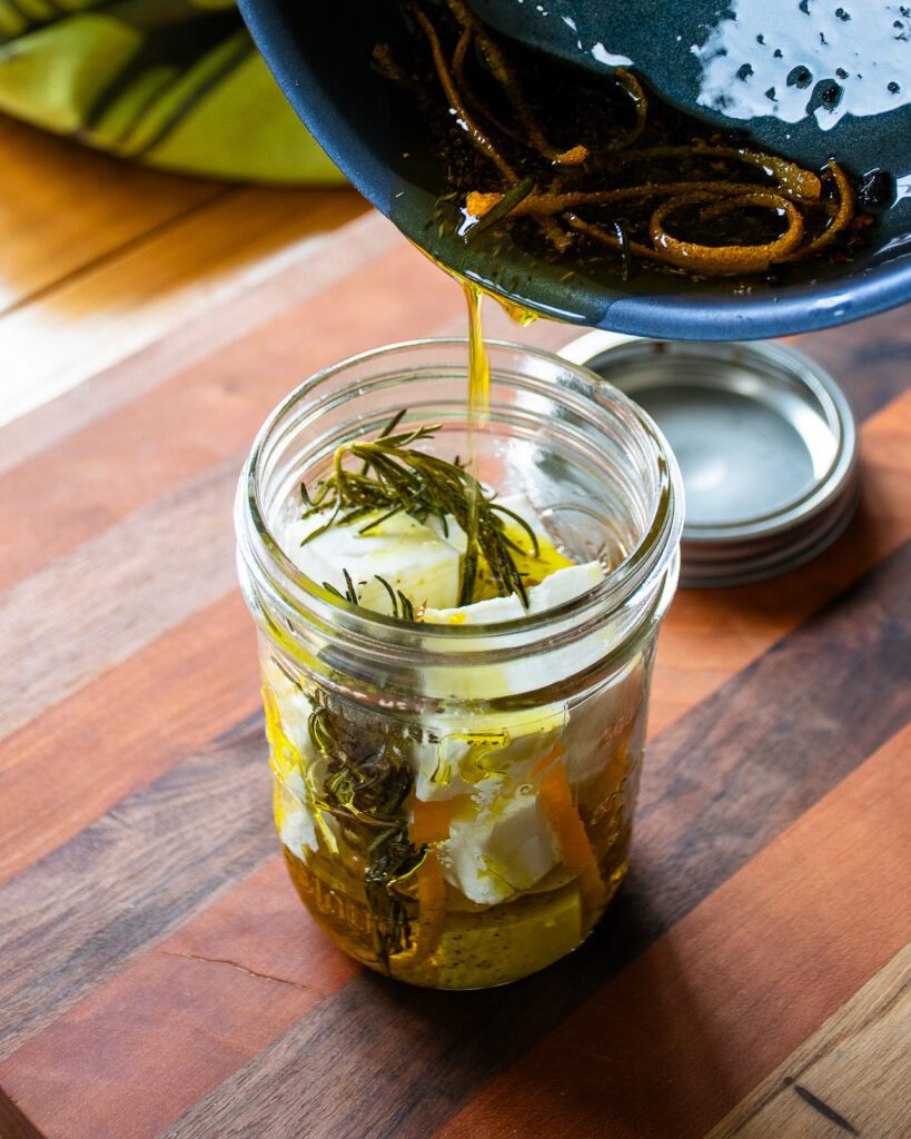 Oil being poured from a skillet into a mason jar with cubed feta inside.