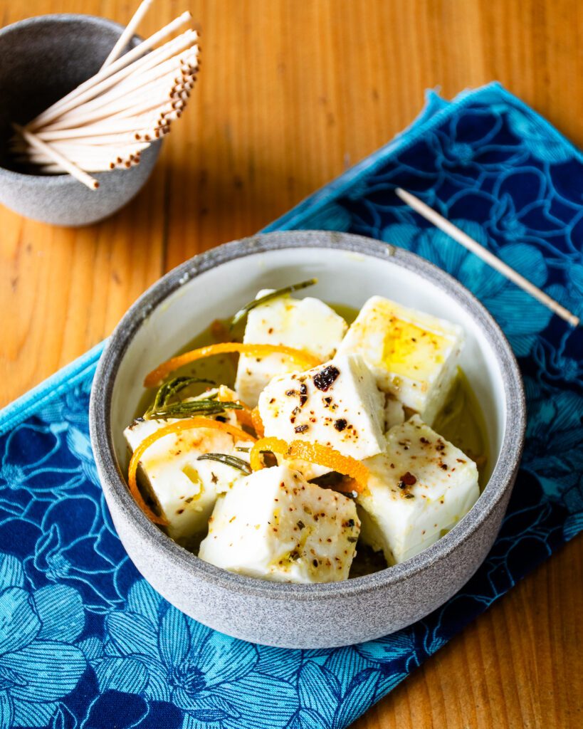 A little dish with marinated feta cheese on a blue napkin with toothpicks near by.