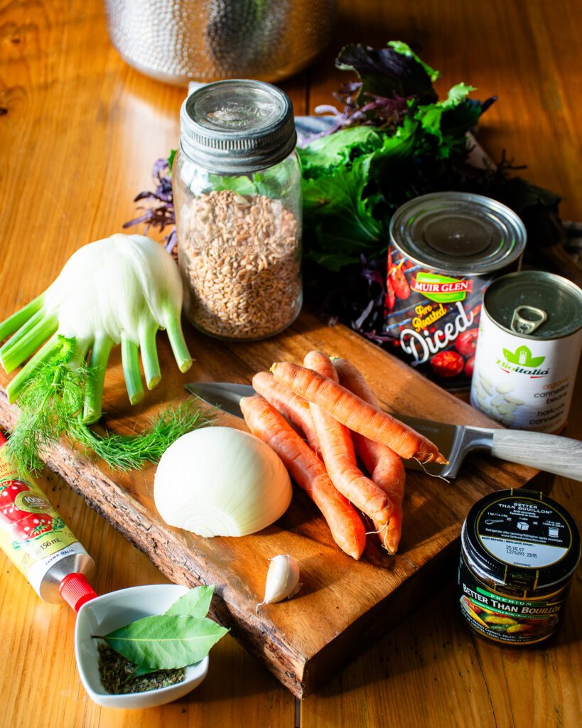 Ingredients on a cutting board on a table - fennel, carrots, onion, herbs and spices, canned tomatoes, canned beans, leafy greens and a jar of farro.