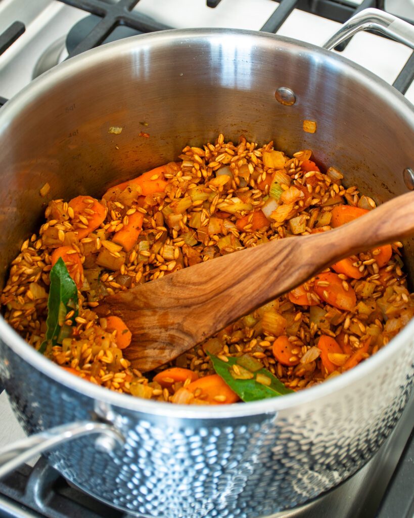 A hammered stainless steel stockpot with vegetables and farro cooking inside and a wooden spoon sticking out of it.