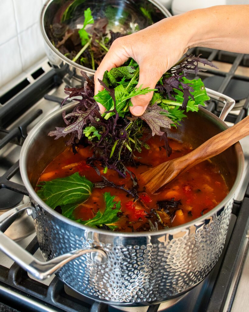 A hand adding leafy greens to a pot of farro soup on the stovetop.