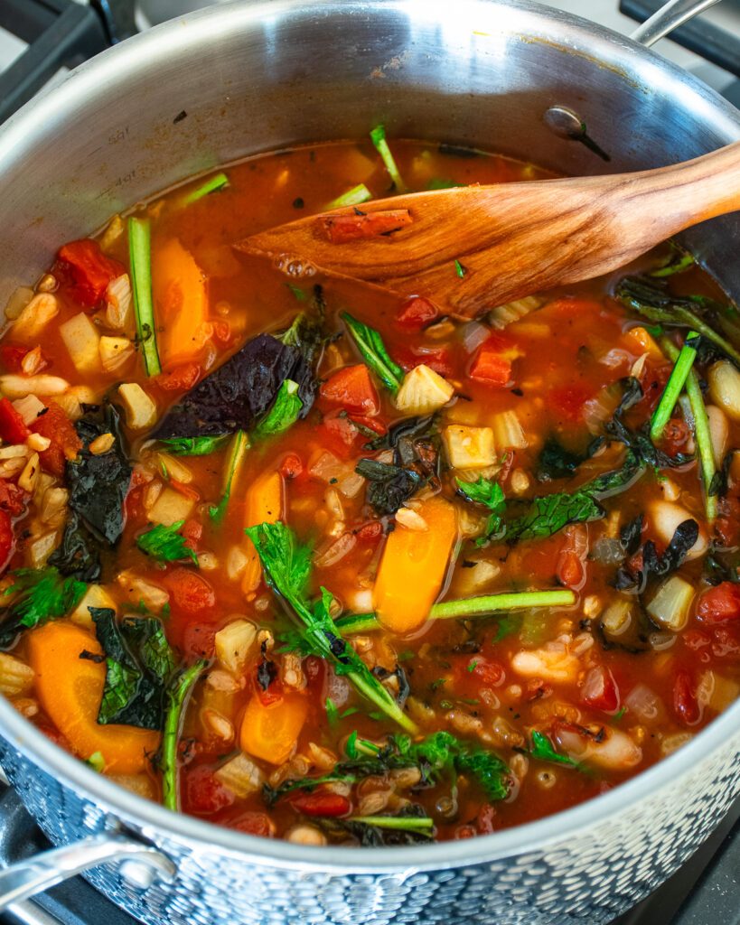 Looking into a stainless steel stockpot with Tuscan farro soup inside and a wooden spoon sticking out.