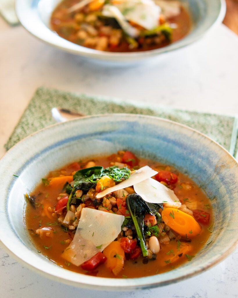 Two bowls of Tuscan farro soup on a white marble table with spoons and napkins.