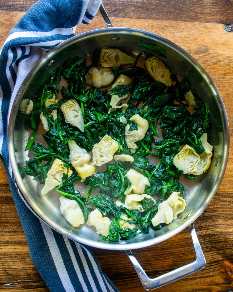 Sautéed spinach and artichokes in a stainless steel saute pan on a wooden background with a blue and white striped towel.