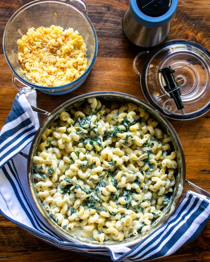 A saute pan with spinach artichoke mac and cheese inside and a chopper of breadcrumbs near by.