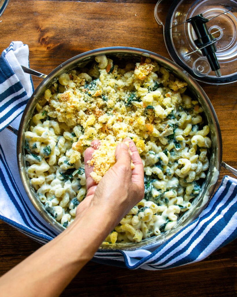 A hand spreading breadcrumbs over a saute pan of mac and cheese.