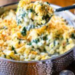 A large serving spoon dishing out some spinach artichoke mac and cheese out of a hammered stainless steel saute pan.