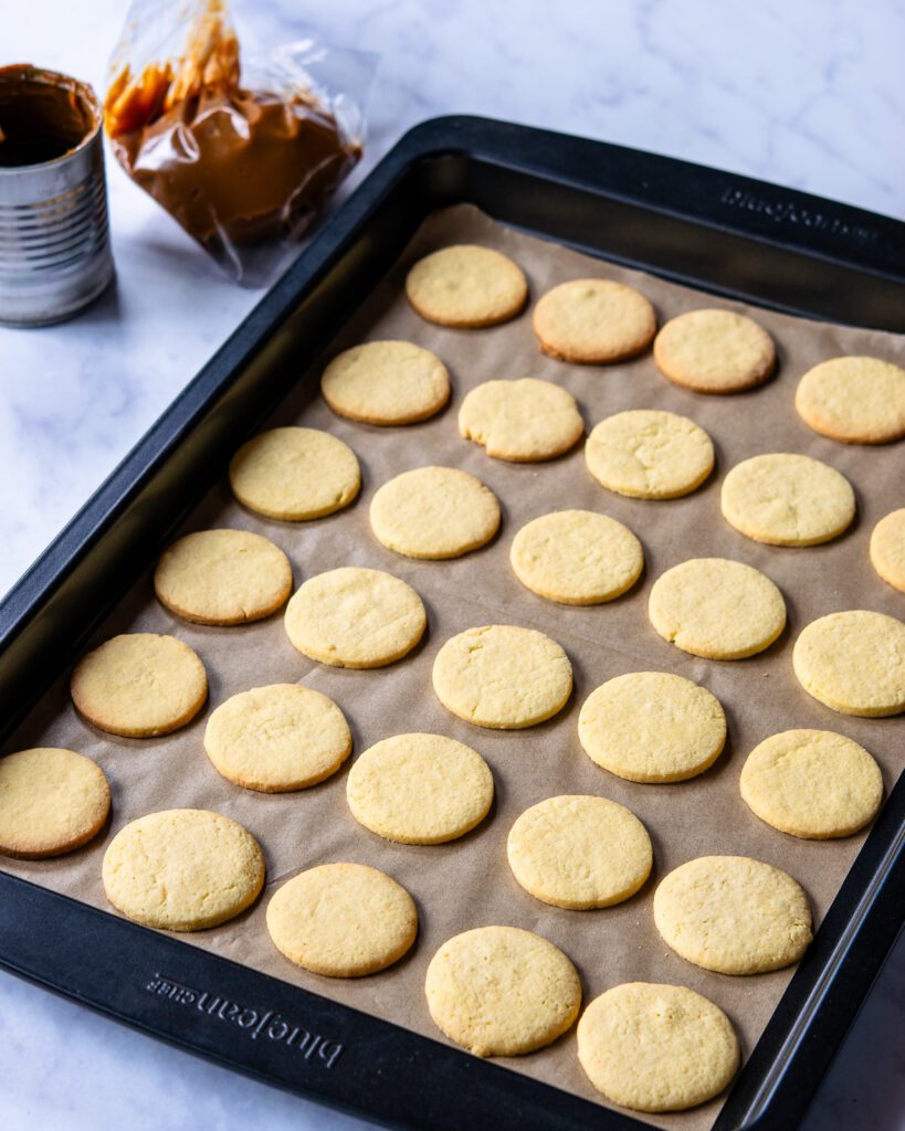 A tray of round butter wafer cookies on parchment paper with a tin of dulce the leche in the background.