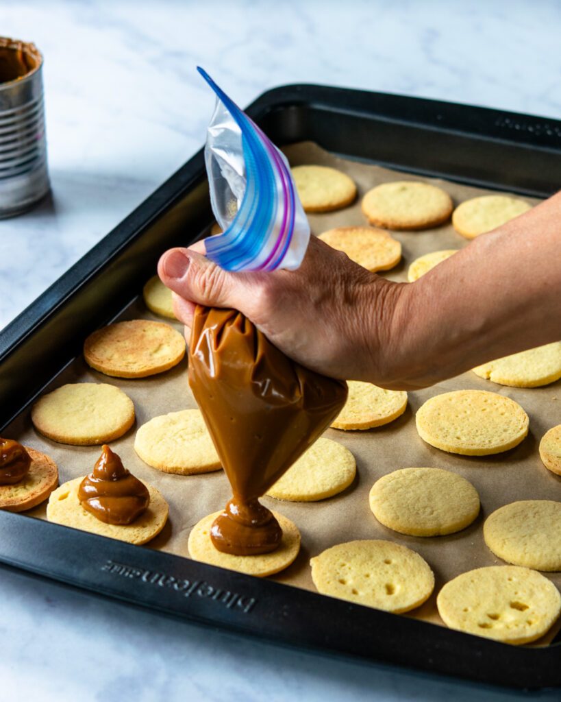 a hand squeezing dulce de leche out of a ziploc bag onto thin butter wafer cookies.