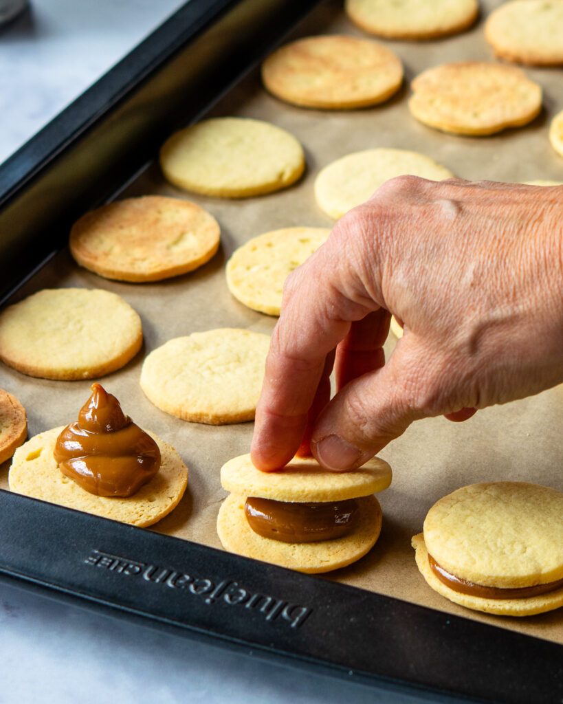 A hand sandwiching two butter cookies with dulce de leche in the middle on a sheet tray.