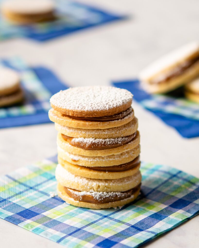 A stack of alfajores on a checkered bev nap with other alfajores in the background.