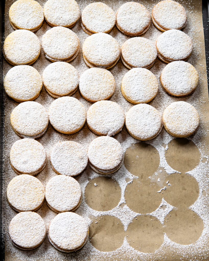 Looking down on a sheet tray with alfajores dusted with powdered sugar and a few spots missing where cookies have been removed.